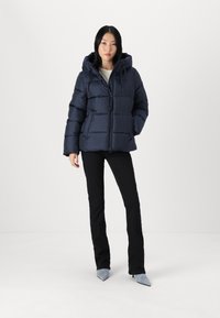 Mornarska puffer jakna s kapuco in z zadrgo. Ima prešite dele, dolge rokave in rahlo sijajno površino. Oblečena je s črnimi hlačami in modrimi čevlji.