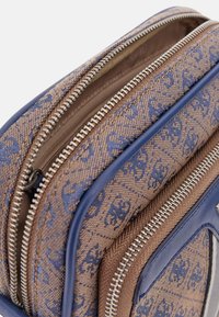Braune Stofftasche mit blauen Akzenten, die eine gemusterte Oberfläche und silberne Reißverschlüsse aufweist. Mehrere Fächer und kontrastierende Texturen.