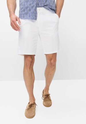 Man met witte knielange shorts, blauw-wit gestreept overhemd en beige veterschoenen, staand tegen een effen witte achtergrond.