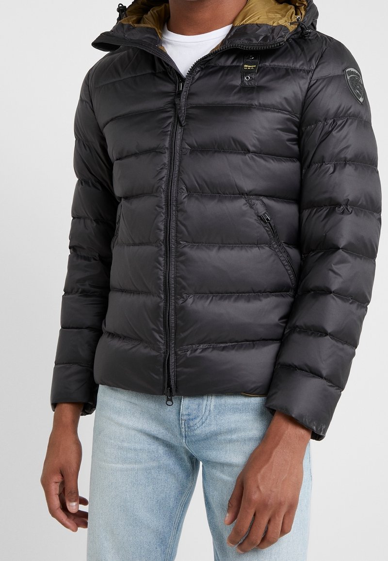 Zwarte pufferjas met een afneembare capuchon, beschikt over een gladde buitenkant, horizontale stiksels en twee ritszakken. Accenten in goud.