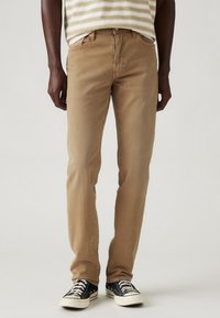 Vaqueros de mezclilla color arena con un ajuste slim, que cuentan con un clásico diseño de cinco bolsillos y un sutil desgaste, combinados con zapatillas de lona en blanco y negro.