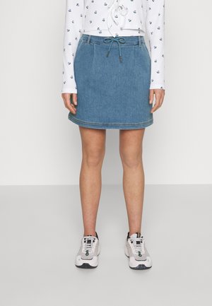 Kobieta ubrana w niebieską dżinsową spódnicę z sznurkiem z przodu, biały top w kwiaty z długim rękawem oraz białe trampki, stojąca na jednolitym tle.