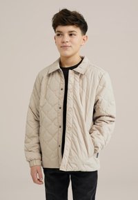 Beige vuorattu takki nappisuljin, terävä kaulus ja sivutaskut, käytetty mustan paidan päällä ja tummaisten housujen kanssa.