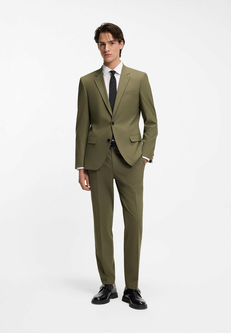 Traje a medida color verde oliva con solapas estructuradas, cierre con un solo botón, pantalones ajustados y zapatos de vestir negros. Camisa blanca y corbata a rayas.