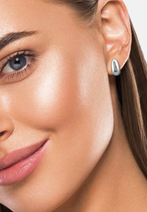 Nahaufnahme eines lächelnden Frauenfaces mit blauem Auge, glatter Haut und einem silbernen, teardrop-förmigen Ohrring im Ohr.