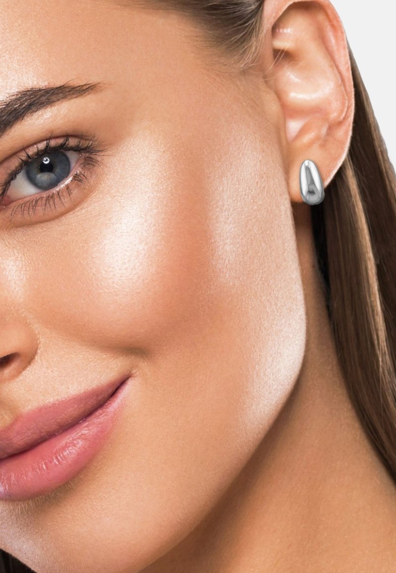 Primo piano del volto sorridente di una donna che mostra un occhio blu, una pelle liscia e un orecchino a forma di goccia d'argento appeso all'orecchio.