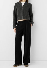 Cardigan gris en maille raccourcie avec fermeture éclair et texture côtelée, associé à un pantalon large noir et des chaussures noires.