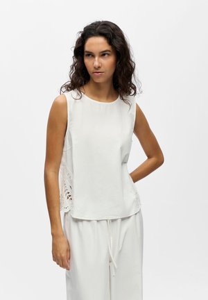 Kobieta z kręconymi ciemnymi włosami nosi białą bezrękawnik z koronkowymi panelami po bokach oraz luźne białe spodnie, stoi z jedną ręką na biodrze.