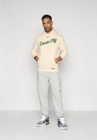 Kremna kapuca s "Cream City" grafiko v zeleni barvi, kombinirana z svetlosivimi cargo trenirkami. Ima kapuco s vrvico in žep.