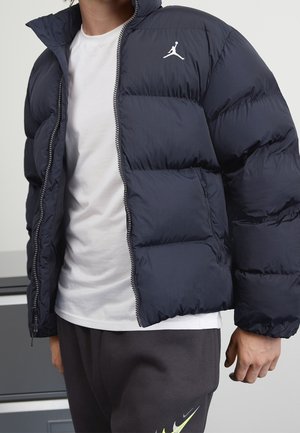 Homme portant une doudoune bleu marine avec un logo Jordan blanc sur la poitrine, un t-shirt blanc et un pantalon noir avec un logo Nike vert néon.