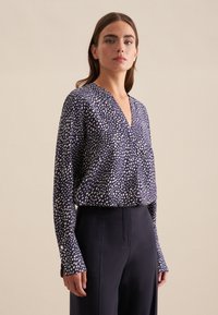 Blusa blu navy con motivo a pois bianchi, scollatura a V, maniche lunghe e dettagli con bottoni ai polsini; abbinata a pantaloni scuri.