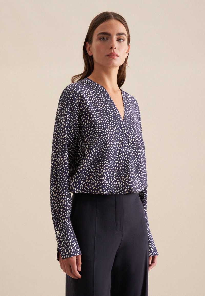 Blusa blu navy con motivo a pois bianchi, scollatura a V, maniche lunghe e dettagli con bottoni ai polsini; abbinata a pantaloni scuri.