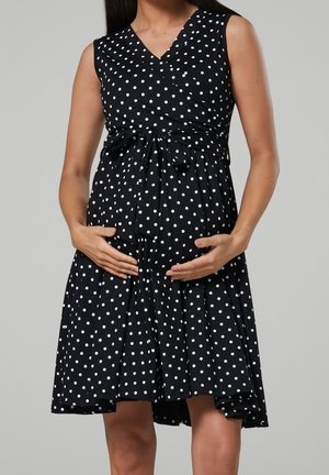 Robe noire sans manches à pois blancs, avec un design croisé à la taille et une jupe évasée arrivant au genou.