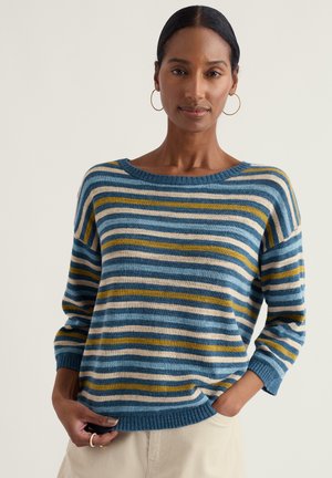 Mujer de cabello oscuro con pendientes de aro dorados y suéter a rayas azules, amarillas y blancas, posando con una mano en el bolsillo y fondo neutro.