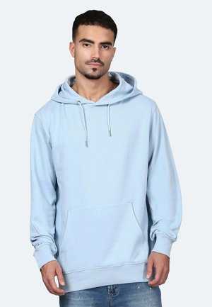 Mann trägt hellblauen Kapuzenpullover mit Fronttasche und blaue Jeans, steht vor einem schlichten hellen Hintergrund.