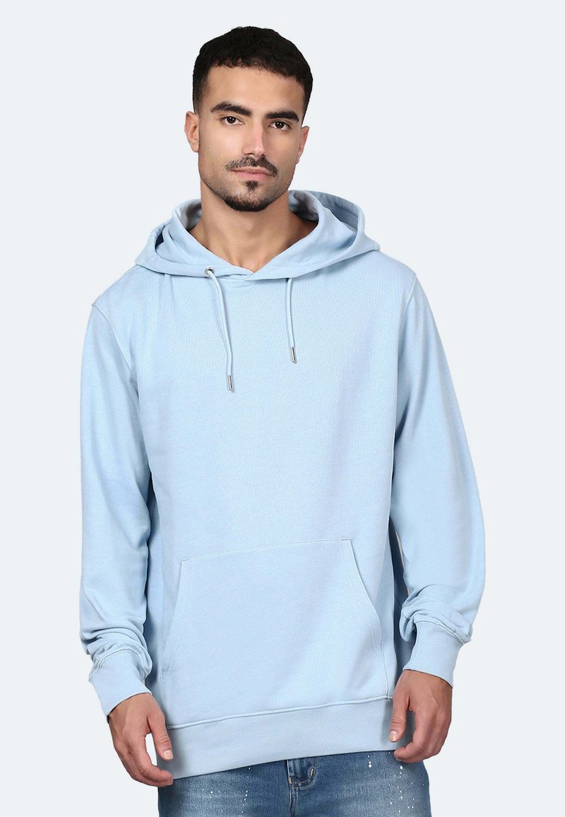Mann trägt hellblauen Kapuzenpullover mit Fronttasche und blaue Jeans, steht vor einem schlichten hellen Hintergrund.