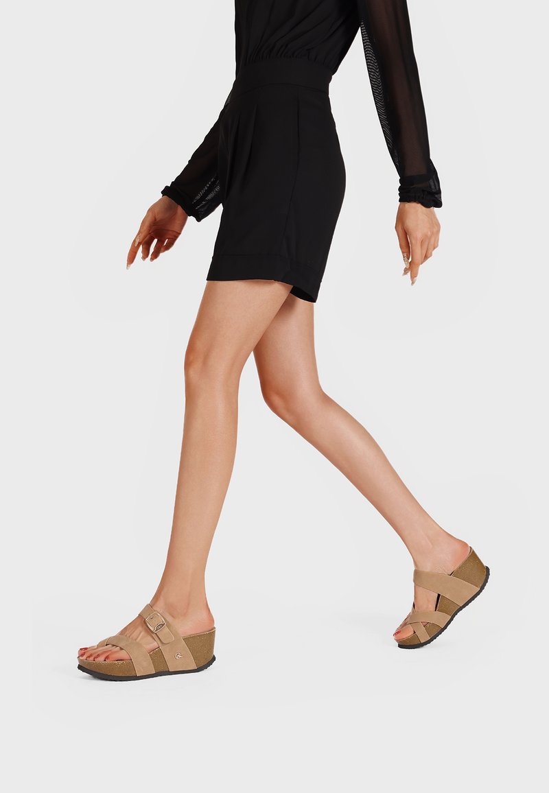 Sandali con zeppa color tan, dotati di due fasce larghe e una fibbia. Sottopiede in sughero con una suola in gomma nera texturizzata. Il modello indossa un abbigliamento nero.