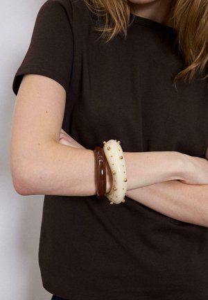 Persona con pelle chiara che indossa una maglietta a maniche corte marrone scuro, con le braccia incrociate, e due braccialetti: uno marrone e uno bianco con borchie dorate.