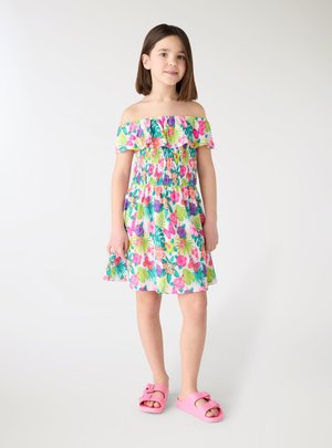 Ragazza giovane in piedi che indossa un vestito floreale senza spalle e sandali rosa su uno sfondo chiaro uniforme.
