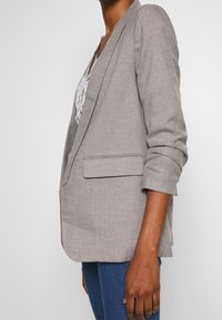 Personne portant un blazer gris clair cintré avec les manches retroussées sur une chemise à motifs et un jean bleu, debout de profil sur fond blanc.