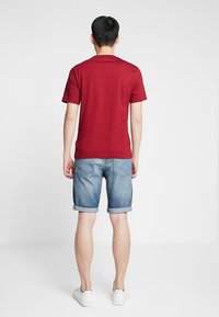 Röd kortärmad skjorta, denimshorts med uppvikta fållar och vita sneakers, sedd bakifrån mot en enfärgad bakgrund.