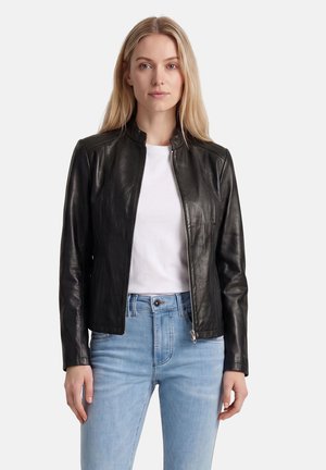 Blonde Frau, die eine schwarze Lederjacke über einem weißen Hemd und hellblauen Jeans trägt, steht vor einem schlichten weißen Hintergrund.