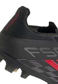 Κοντινό πλάνο μαύρου ποδοσφαιρικού παπουτσιού με κόκκινο λογότυπο "F50" και υφασμένο συνθετικό επάνω μέρος, εμφανίζοντας τις τάπες και την περιοχή της φτέρνας.