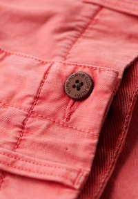 La tela de color coral presenta costuras visibles, una superficie texturizada y un botón marrón oscuro con "SUPERDRY" en relieve.