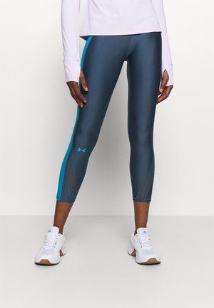 Jambes portant des leggings bleu foncé avec des rayures bleu sarcelle et des panneaux en mesh, assortis à des baskets blanches, sur un fond clair uni.