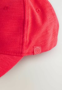Gorra roja hecha de tela texturizada con detalles de costura. Presenta una pequeña etiqueta con una carita sonriente. La visera se extiende hacia adelante.