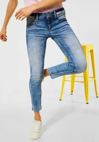Modré džíny s úzkým střihem, které mají vybledlý efekt, přední kapsy a střední výšku pásku. V kombinaci s bílými teniskami na pozadí žlutého stoličky.