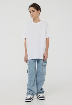 T-shirt bianco oversize, jeans blu a taglio ampio con tasche laterali, indossati con sneakers bianche e nere. Outfit semplice e casual.