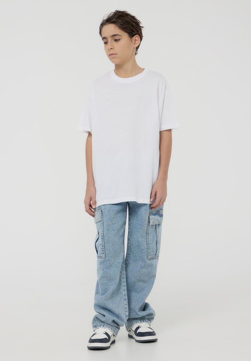 Biała, oversize'owa koszulka, niebieskie luźne dżinsy z kieszeniami bocznymi, zestawione z czarno-białymi trampkami. Prosty, casualowy strój.