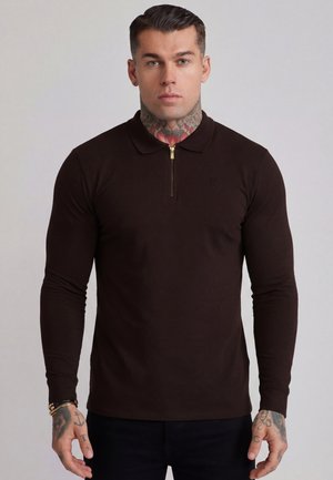 Mand med tatoveringer på hals og hænder iført en tætsiddende langærmet mørkebrun polo med en gylden lynlås, stående mod en ensfarvet baggrund.
