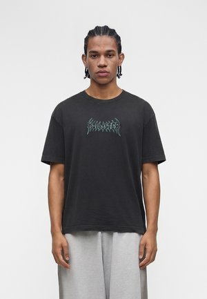 Νεαρός άνδρας με πλεξούδες, φοράει φαρδιά μαύρη μπλούζα με πράσινο γραφικό λογότυπο και ανοιχτό γκρι παντελόνι, στέκεται μπροστά σε απλό φόντο.