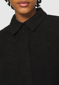 Chemise en flanelle noire avec col, tissu texturé, cinq boutons et fermeture à boutons sur le devant. Vue rapprochée mettant en avant le matériau et le design.