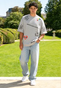 Jeune homme marchant en plein air portant un t-shirt gris surdimensionné avec du texte et des graphismes imprimés, un jean bleu clair et des baskets blanches.