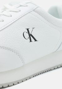 Bílé kožené tenisky s texturovaným svrškem, které mají černé logo "ck", bílé šňůrky a světlou šedou gumovou podrážku s ribbed vzorem.