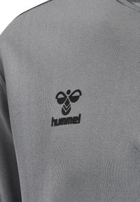 Nahaufnahme eines hellgrauen Stoffes mit einem kleinen schwarzen hummel-Logo, das eine stilisierte Hummel und Text im Brustbereich zeigt.