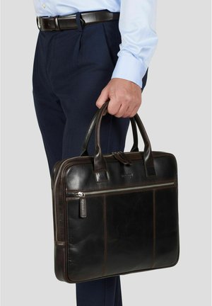 Mann i blå skjorte og marineblå bukser holder en mørkebrun lærmappe med doble håndtak og en glidelåslomme foran.