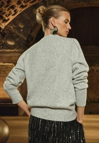 Šviesiai pilkas megztinis su ilgomis rankovėmis ir laisvu siluetu, derintas su juoda fringinė sijonu. Apskriti auskarai suteikia išbaigtumo visam įvaizdžiui.