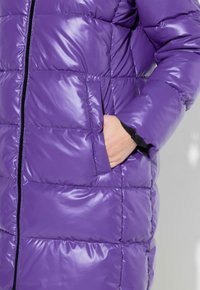 Chaqueta de abrigo morada con un acabado brillante, que presenta patrones de acolchado diagonales, un bolsillo lateral y un detalle de cremallera negra.