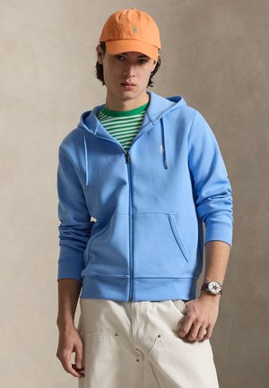 Mladý muž nosí oranžovú čiapku, zelenú a bielu pruhovanú košeľu, modrú mikinu so zipsom, béžové nohavice a náramkové hodinky s farebným ciferníkom.