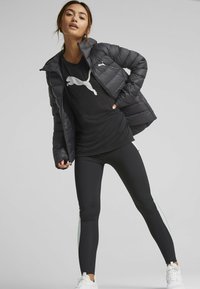 Sort pufferjakke med en blank finish, parret med en sort T-shirt med et hvidt logo, og sorte leggings med lysegrå sidelister. Hvide sneakers.