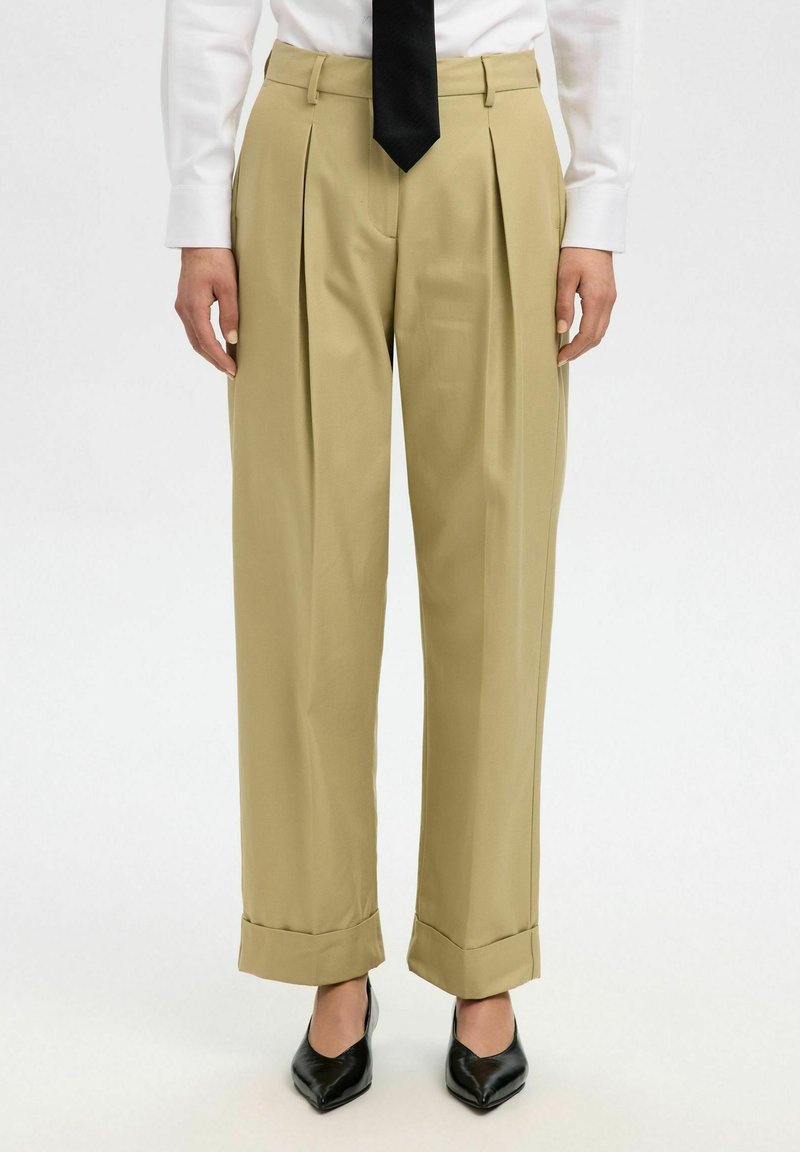 Beige Plissee-Hosen mit hoher Taille und weitem Bein, mit umgeschlagenem Saum, die über schwarzen spitz zulaufenden Schuhen sitzen.