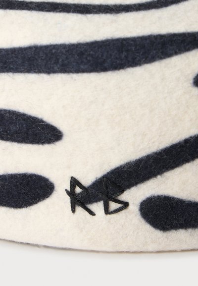 Gros plan d'un tissu crème avec des formes abstraites noires et des lettres brodées noires "RB" près du bord inférieur.