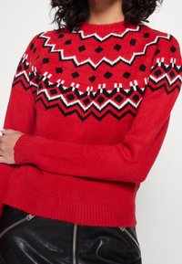Czerwony sweter wykonany na drutach z geometrycznym wzorem w białym i czarnym kolorze, z okrągłym dekoltem oraz ściągaczami przy mankietach i u dołu.