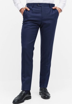 Mand iført marineblå skræddersyede bukser, hvid skjorte stoppet ned i bukserne, sorte læder loafers, stående med hænderne i lommerne på hvid baggrund.