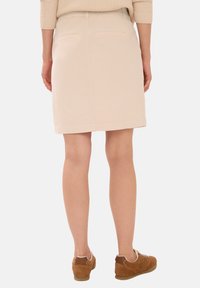 Žena v béžové sukně ke kolenům, s hnědými teniskami a světle hnědým svetrem, stojící zády k bílé pozadí.