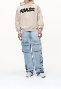 Persona vestida con sudadera beige con texto negro, pantalones cargo de mezclilla azul claro con múltiples bolsillos y zapatillas blancas sobre un fondo blanco.